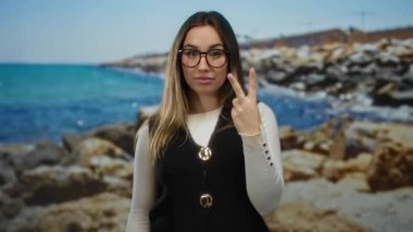 Güneşli bir plajda neşeli bir ifadeyle kutluyor, sarışın, gözlüklü, sakin deniz manzarasının ortasında heyecanlı görünen bir kadın..