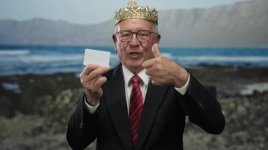 Sahilde takım elbiseli kıdemli bir adam, elinde bir kartla arka planda dalgaların çarptığı bir taç tutarken başparmağını kaldırıyor..