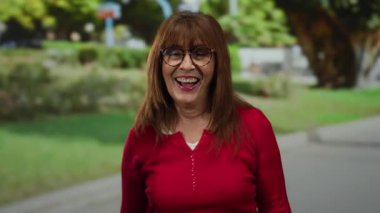 Kırmızı gömlekli yaşlı bir kadın, canlı bir parkta gülümserken, taştan jest yapıyor. Yeşillik ve görünür yürüyüş yolu var, neşe ve coşku saçıyor..