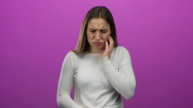 Yanağını acıyla bastıran genç bir kadın canlı pembe bir arka plana karşı duruyor, beyaz bir kazak giyiyor, sıkıntısını ve rahatsızlığını ifade ediyor, yalnız ve düşünceli..