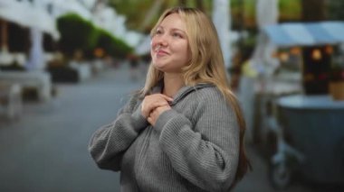 Şehir sokaklarında gülümseyen kadın gri kazak giyiyor, neşe ve özgürlüğü somutlaştırıyor, yeşillik ve sokak satıcıları arka planda bulanıklaştığı için şehir ortamında gevşek davranışlar sergiliyor..