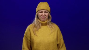 Sarı bir süveter ve şapkayla, parlak mavi bir arka planda, tiksinmiş bir ifade sergileyen bir kadın..