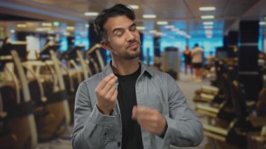 Genç İspanyol bir adam spor salonunda, spor aletleri ve tavan lambalarıyla çevrili bir şekilde para jesti yapıyor. Bu da spor ortamında motivasyon ya da finansal hırs olduğunu gösteriyor..