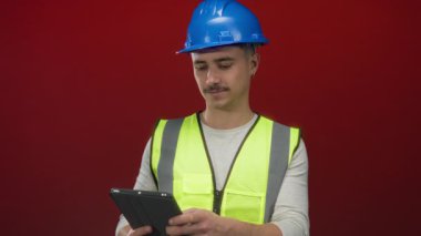 Yansıtıcı yelek ve şapka giyen genç İspanyol adam izole edilmiş kırmızı arka plan duvarına karşı bir tablet kullanıyor.