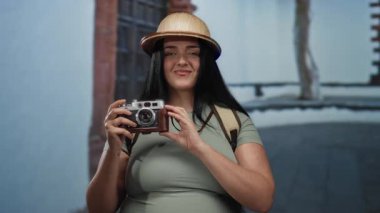 Kamera ve sırt çantalı bir kadın turist gülümseyerek şehir sokaklarını keşfediyor, canlı bir şehir ortamında açık hava maceralarını yakalıyor..
