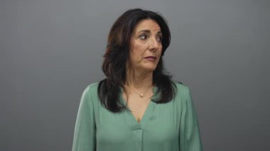 Orta yaşlı İspanyol kadın gri stüdyoda el kaldırıyor yan gözle bakıyor ve el hareketlerini sergiliyor..