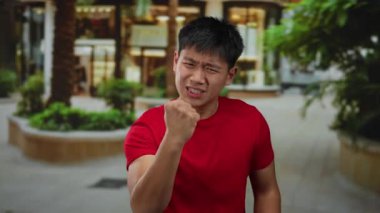 Kırmızı gömlekli genç adam şehir caddesinde hayal kırıklığı yaşıyor. Çevresi bulanık yeşillik ve şehir mimarisi ile çevrili. Açık hava duygularının özünü yakalıyor..