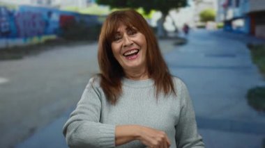 Bir şehir caddesinde gülümseyen ve avucuna işaret eden İspanyol son sınıf öğrencisi bir kadın gri bir kazak giyiyor, arka planda renkli bir grafiti var, açık hava ortamının keyfini çıkarıyor..