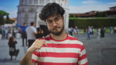 Güneşli bir günde sidik kulesinin önüne işaret eden gözlüklü ve sakallı İspanyol genç adam İtalya 'da turist ve mimariyle çevrili..