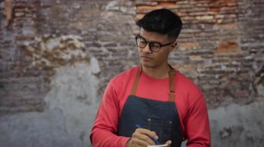 Gözlüklü ve önlüklü genç İspanyol bir adam şehir sokaklarında notlar yazıyor, kentsel çevre ile etkileşime girerken dikkatle odaklanıyor..