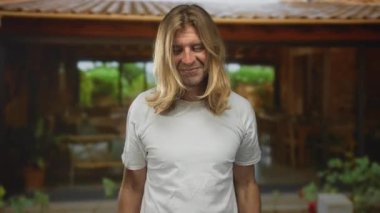 Beyaz tişörtlü, uzun saçlı sarışın adam bina girişinde kaya boynuzları hareketi yapıyor..