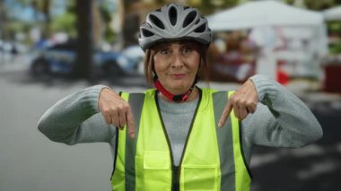 Şehir sokaklarında kıdemli İspanyol bir kadın kask ve yansıtıcı yelek giyiyor, elleri aşağıda, kalabalık bir şehir ortamının ortasında gülümsüyor..