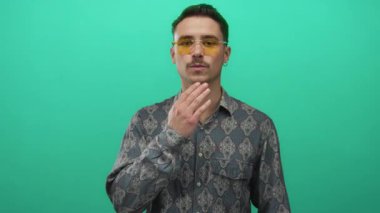 Şablonlu gömlekli ve sarı gözlüklü genç adam canlı yeşil arka planda duruyor. Kendine güvenen ve şık görünüyor..