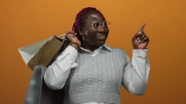 Alışveriş torbalarıyla gülümseyen kadın, izole edilmiş turuncu bir arka plana sevinçle işaret ediyor. Bu da olumlu bir alışveriş deneyimi ve coşku olduğunu gösteriyor..