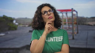 Yeşil tişörtlü bir kadın arka planda spor aletleriyle dışarıda düşünüyor..