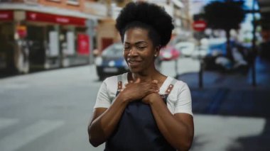 Önlük giymiş bir kadın, bulutlu bir günde şehir caddesinde elleri göğsünde, duygularını ifade ederek araba ve binaların önünde dikiliyor..