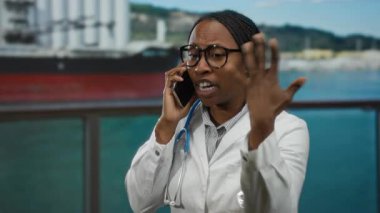 Deniz kenarı limanı yakınlarında akıllı telefondan konuşan bir kadın. Gözlük takıyor, steteskop takıyor ve açık havada bir tekne kullanıyor..
