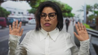 Şehir caddesinde dikilen kadın, elleri havada, gözlüklü ve beyaz gömlekli, artı boyutu Latin, İspanyol güzelinin şehir ortamında yakalandığı şüphesini dile getiriyor..