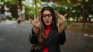 Gözlüklü genç bir kadın, her iki orta parmağıyla bir jest yapıyor. Açık havada bir parkta gülümserken. Etrafı yeşillik ve sonbahar yapraklarıyla çevrili..