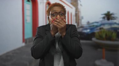 Genç Afro-Amerikalı kadın, güneşli caddede binanın girişinde elleri ile ağzını kapatıyor. Gözlük ve altın küpeler takıyor. Sürpriz..