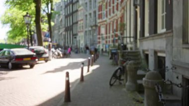 Arka planda Hollanda 'nın Amsterdam kentindeki kaldırım taşı patikası boyunca bulanık yayaları ve park halindeki arabaları gösteren şehir manzarası..