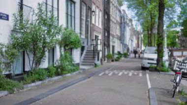 Bulanık Amsterdam caddesi manzarası, çözünmüş binalar, yeşil bitkiler, beyaz bir minibüs, ve mavi gökyüzü altındaki uzak figürler, canlı bir Avrupa kentinin özünü yakalıyor..