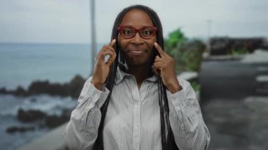 Gözlüklü kadın, deniz kenarında bir gezinti güvertesinde neşeli bir şekilde gülümsüyor. Okyanusun sakin arka planına karşı enerjik bir enerji ve ifade sergiliyor..