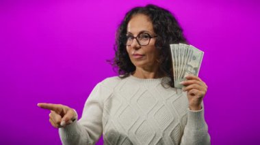 Gözlük ve süveter takan, kendine güveni tam bir mali başarı ifadesiyle işaret eden, izole edilmiş mor bir arka plan karşısında Amerikan doları tutan orta yaşlı bir kadın..