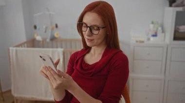 Yatak odasında akıllı telefon kullanan bir kadın bebek beşiğinin yanında oturuyor, kızıl saçlı, gözlüklü, kırmızı kazaklı, modern ev içi, huzurlu bir atmosfer..