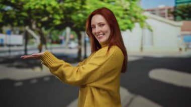 Güneşli bir şehir caddesinde, ağaçlar ve binalarla dolu sarı bir kazakla gülümseyen bir kadın, şehir atmosferiyle neşeli bir açık hava sahnesi yaratıyor..