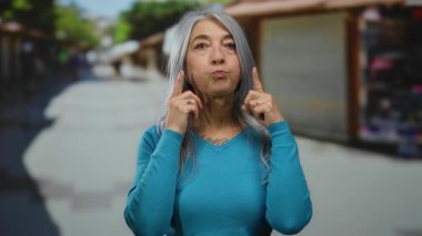 Gri saçlı, mavi kazaklı son sınıf öğrencisi bir kadın açık hava pazarında duruyor ve parlak bir sokak ortamında arka planda neşeli bir yüz ifadesi sergiliyor..