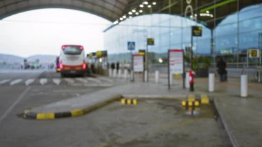 Açık hava havaalanı terminalinin bulanık görüntüsü. Odaklanmış otobüs ve yolcular bulanık bir kaldırım üzerindeki bokeh ışıklarını yansıtıyor. Belirsiz bir seyahat atmosferi yaratıyor..