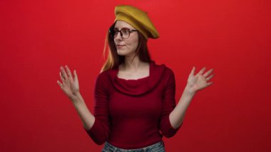 Kızıl saçlı, gözlüklü ve sarı bereli bir kadın canlı kırmızı arka plan duvarının önünde el ele tutuşarak poz veriyor..