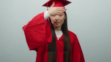 Kırmızı mezuniyet elbisesi giymiş, yüzü elle kaplanmış bir kadın beyaz bir arka plana karşı poz veriyor.