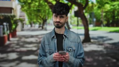 Şehirdeki sokak ortamında telefonu olan genç adam, biraz hoşnutsuz görünüyor, arka planda ağaçlar ve binalar olan kot ceket giyiyor güneşli bir günde..