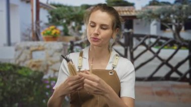 Bir evin yakınındaki bahçede fırça tutan kadın ressam önlük giyiyor ve düşünceli bir ifadeyle sanatsal aletlerine odaklanıyor..