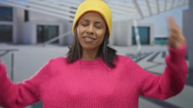 Pembe kazaklı ve sarı bereli genç İspanyol kadın şehir ortamında gölgeler ve modern mimariyle neşeli bir ifade sergiliyor.