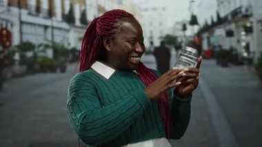 Dolarlarla dolu kavanozu elinde tutan kadın açık havada gülümsüyor para biriktirerek mutluluğunu gösteriyor Afro-Amerikan kadın şehir ortamında finans zevkini çıkarıyor.