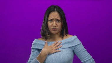 Göğsünde eli olan genç bir kadın, açık mavi bir kazak giyen, canlı bir mor arka plan karşısında dururken şaşırmış görünüyor. İnançsızlık ve duyguyu ifade ediyor.