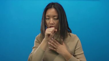 Asyalı bir kadının mavi arka planda öksürmesi, sağlık sorunlarını ve rahatsızlıkları vurguluyor.