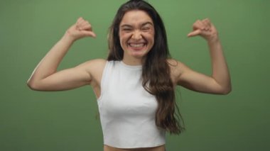 Yeşil bir stüdyoda gülümseyen ve başparmağıyla göğsünü gösteren Latin asıllı genç bir kadın..