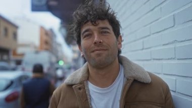 Kentsel bir sokak ortamında İspanyol bir adam sıcacık gülümsüyor. Tuğla duvara yaslanmış kahverengi ceketli sıradan bir tarz sergiliyor. Şehir hayatı ve park halindeki arabalarla çevrili..
