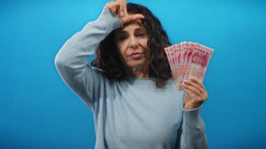 Orta yaşlı bir kadın, Çin Yuan banknotlarını mavi arka planda tutarak, duvarın içeriğinden izole edilmiş, kültürel ve finansal temalar yakalayan, kaybeden tabelalarını mizahi bir şekilde ifade ediyor..