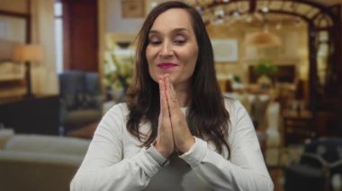 Lüks bir otel salonunda, gözleri kapalı meditasyon yapan, beyaz bir gömlek giyen, zarif bir iç mekan ortamında sakinlik ve rahatlık saçan bir kadın..