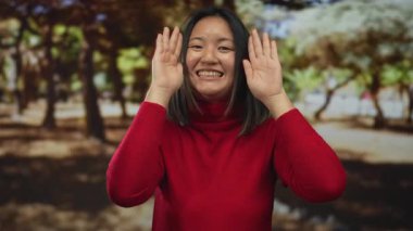 Kırmızı kazaklı genç kadın, neşeli ağaçlar ve doğal güzelliklerle çevrili güneşli bir parkta gülümseyen yüzünü gösteriyor..