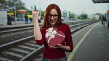 Tren istasyonundaki kızıl saçlı kadın, taş gibi hareketler yaparken gülümsüyor, kalp şeklinde hediye kutusunu açık havada tutuyor, gözlük ve kırmızı kazak giyiyor, neşeli bir anı yakalıyor..
