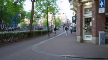 Bokeh etkisi olan Avrupa 'nın bir caddesinde yürüyen ve bisiklet süren insanların bulanık görüntüsü, gün ışığında ağaçların ve dükkanların bulunduğu açık hava kentsel manzarasını gösteriyor.