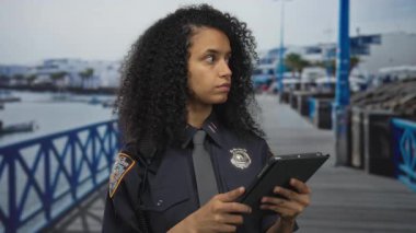 Tabletli kadın polis, deniz kenarındaki gezinti alanında rozetli bir kadın polis memuru, açık deniz kıyısında şehir manzarasında görev yapıyor..