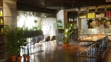 Bitkiler, masalar ve sandalyelerle dolu modern bir restoranın bulanık görüntüsü, yumuşak ışıklandırma ve kentsel yemek tarzını yakalayan çözülmüş elementler aracılığıyla sakin bir atmosfer yaratıyor..