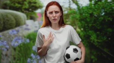Parkta kızıl saçlı bir kadın elinde bir futbol topu tutarken yeşil ve canlı çiçeklerle çevrili beyaz bir gömlek, şaşkınlık ve şaşkınlık ifade ediyor..